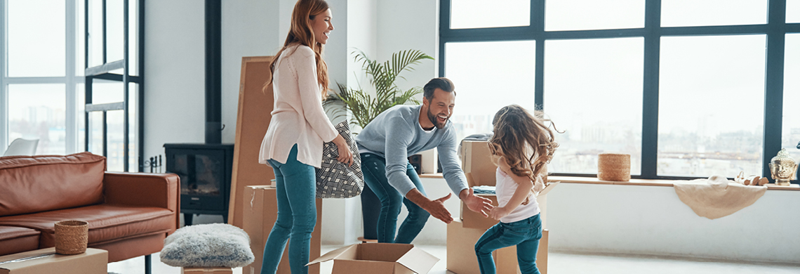 Drei Personen in einem modernen Wohnzimmer mit großen Fenstern, die Umzugskartons auspacken; eine Person hält einen Korb, eine andere hilft einem Kind aus einem Karton.